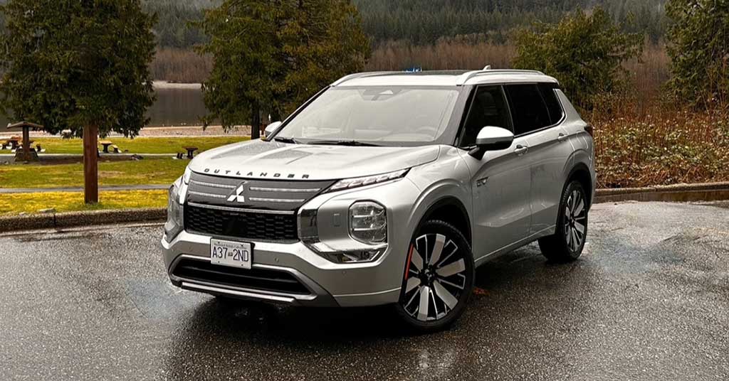 2026-Outlander-PHEV - Two Hills Voice Front-three-quarter view of a silver Mitsubishi Outlander SUV parked on a wet pavement with trees and a lake in the background, daytime outdoor scene.