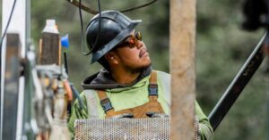 Construction worker in a hard hat and safety glasses looks up at a task beside scaffolding and mesh panels outdoors.